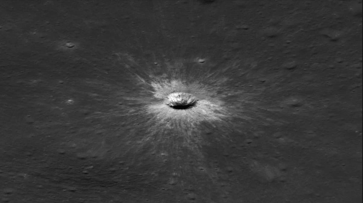 An aerial view of a lunar crater. A bright, circular impact crater sits in the center of a dark, cratered surface, with white rays of ejected lunar material radiating outwards in all directions, creating a stark contrast against the gray terrain.
