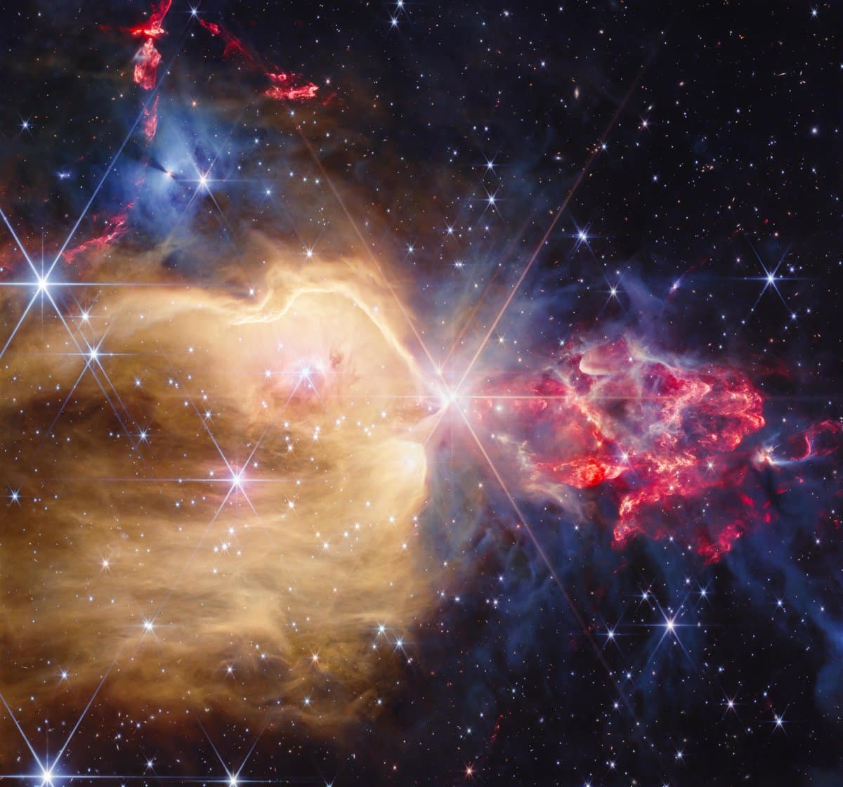 A young star cluster filled with many stars that display Webb’s unique eight-pronged diffraction pattern. At its center is a yellow star which sports the largest diffraction pattern. To the left of the star, there is a region filled with yellow dust and gas that extends from the star to the left border of the image. This yellow region extends from the bottom, covering about two-thirds of the image. The yellow region has several blue stars of different sizes embedded within. The top third, which lies outside of the yellow region, holds a few bright protostars, within dense gray gas. Those stars illuminate the gas, making it appear blue. A few red outflows from other protostars are visible above this grayish-blue region. To the right of the central star, there is a clumpy, flame-like plume of red dust and gas with protostars that emit a soft white light within. The plume is surrounded by more dense gray gas and dust. 

Image credit: NASA, ESA, CSA, STScI, Processing: Alyssa Pagan.