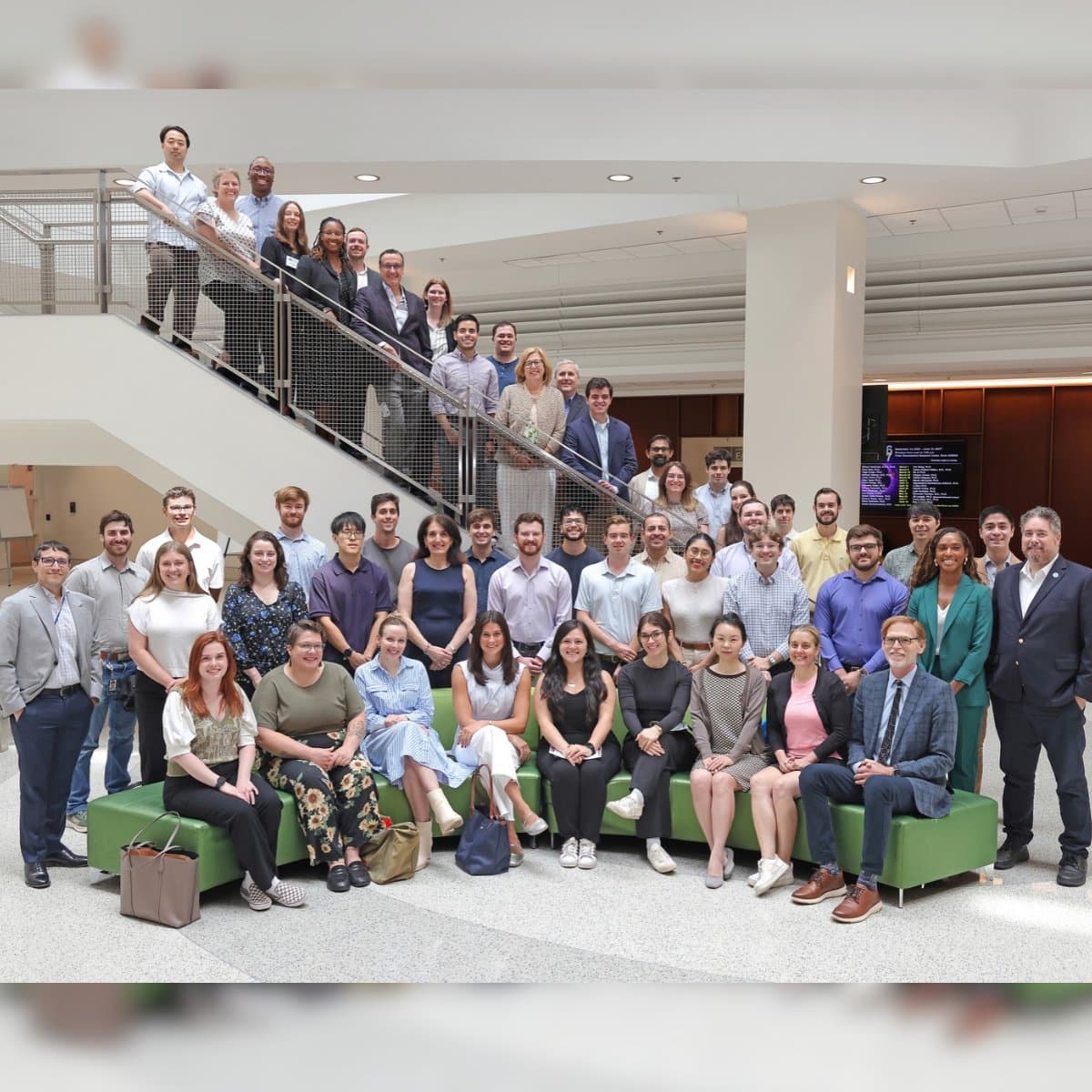 A large group of people posed together in a spacious indoor setting, with some sitting on green couches and others standing on a staircase and nearby. The background includes modern architectural elements and a digital screen displaying text.