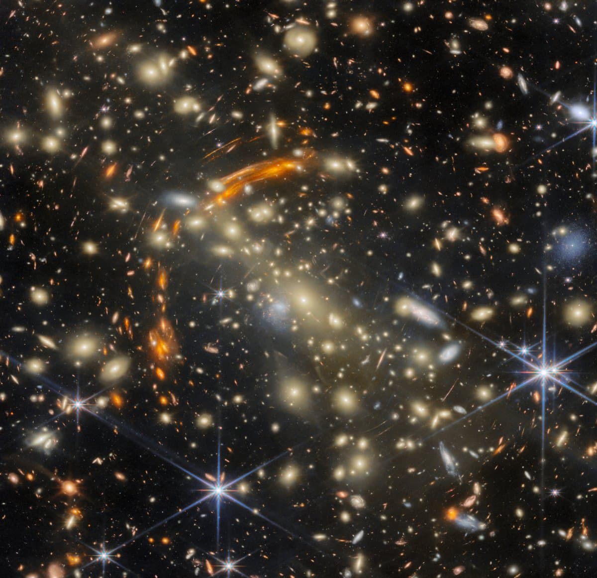 A massive galaxy cluster, made of many golden elliptical galaxies with the largest, brightest one in the center. Heavily distorted orange galaxies appear all around the cluster, the largest concentrated in a large arc around the cluster's center; these are images of very distant galaxies, created by gravitational lensing. A few galaxies are large and bluish-white in color; bright, nearby stars are the same color.
