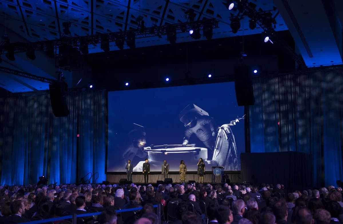 Broadway performers sing on a stage in front of a large crowd during the opening ceremony of the 70th International Astronautical Congress. There is a large screen behind them.
