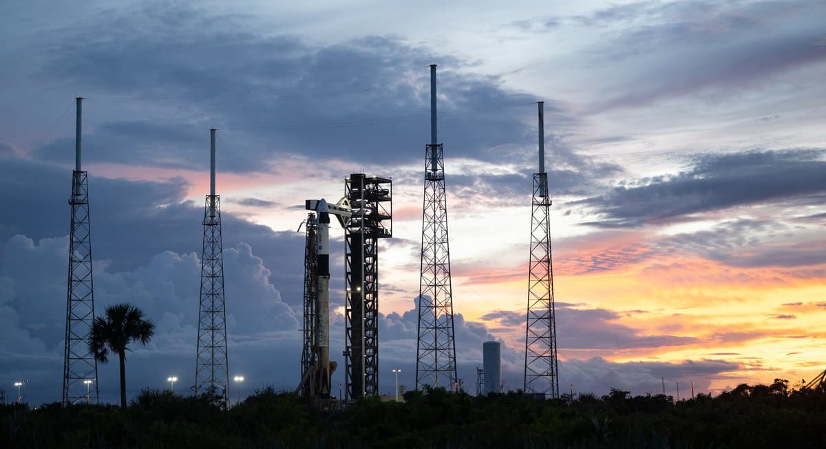 A SpaceX Falcon 9 rocket with the company's Dragon spacecraft on top is seen at sunset on the launch pad. The sky is mostly covered with different kinds of clouds: thick, fluffy ones at the bottom and wider, more spread out ones in the rest of the sky. The Sun's light turns the bottom right portion of the sky orange.