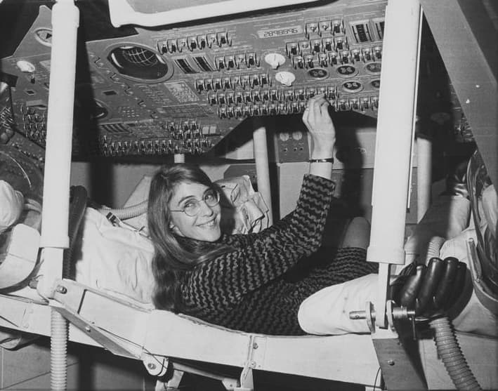 Margaret Hamilton is reclined inside a NASA spacecraft module, surrounded by control panels and switches.